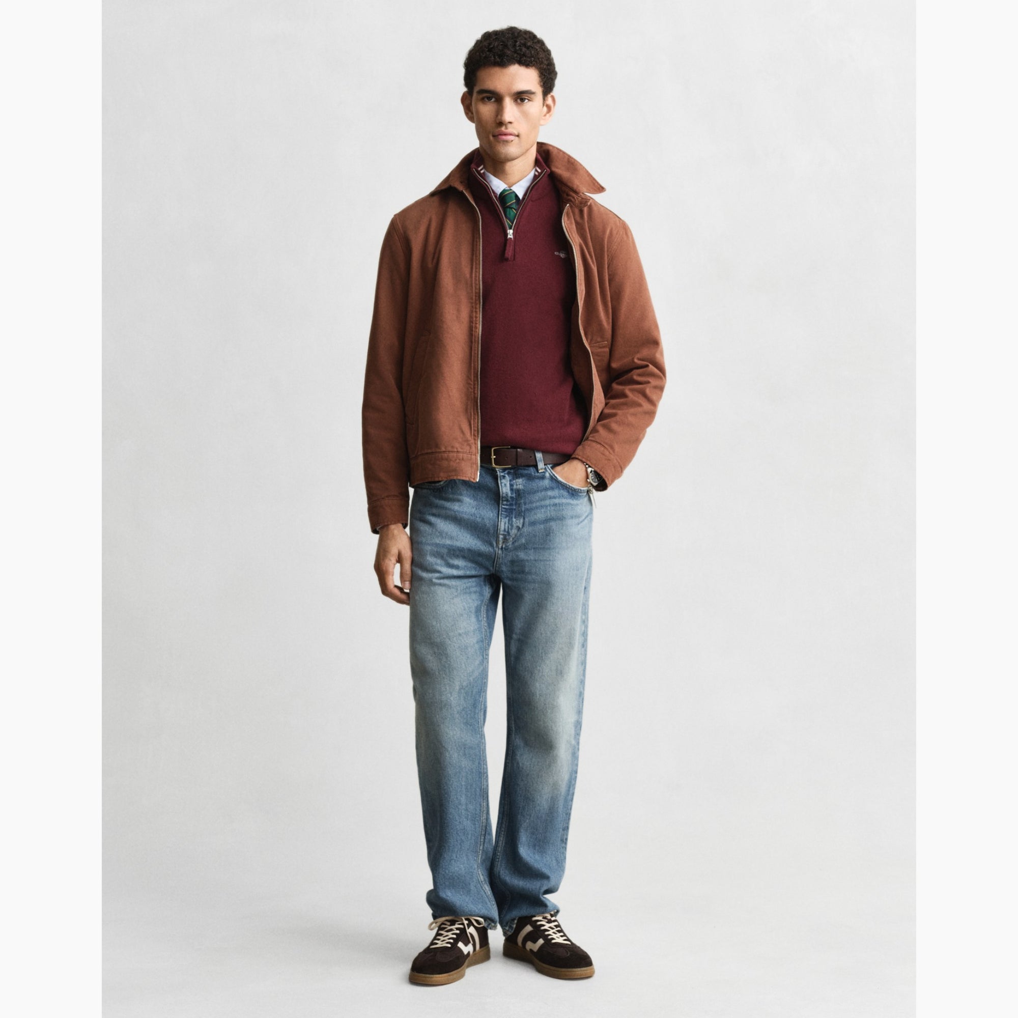 Man wearing a brown GANT jacket, red shirt, blue jeans, and brown shoes on a white background