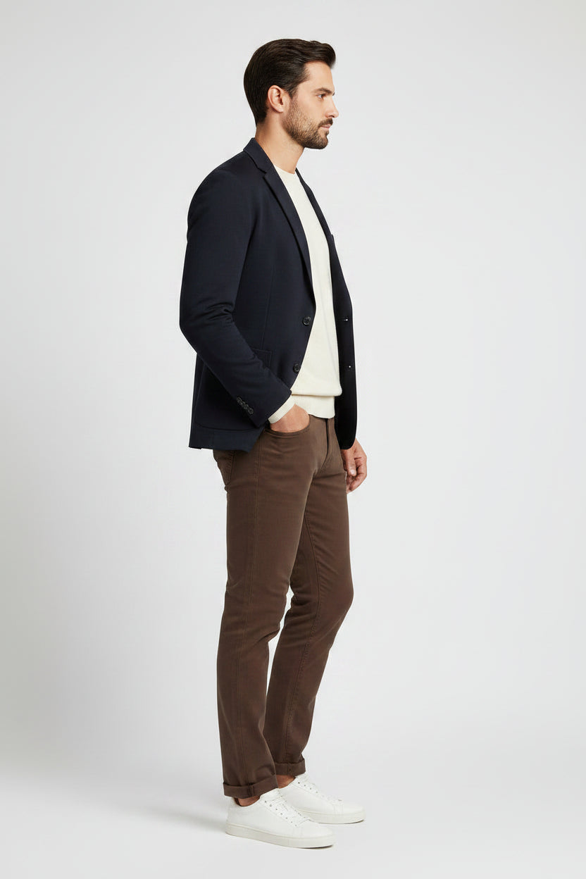 A man modeling a dark navy blazer, cream crewneck sweater, brax brown jeans, and white sneakers against a light gray studio background.