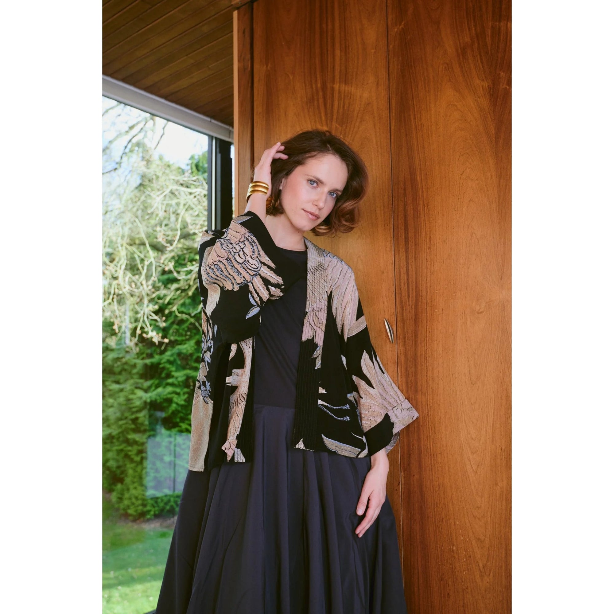 Woman wearing a patterned cardigan over a dark dress, standing against a wooden wall.