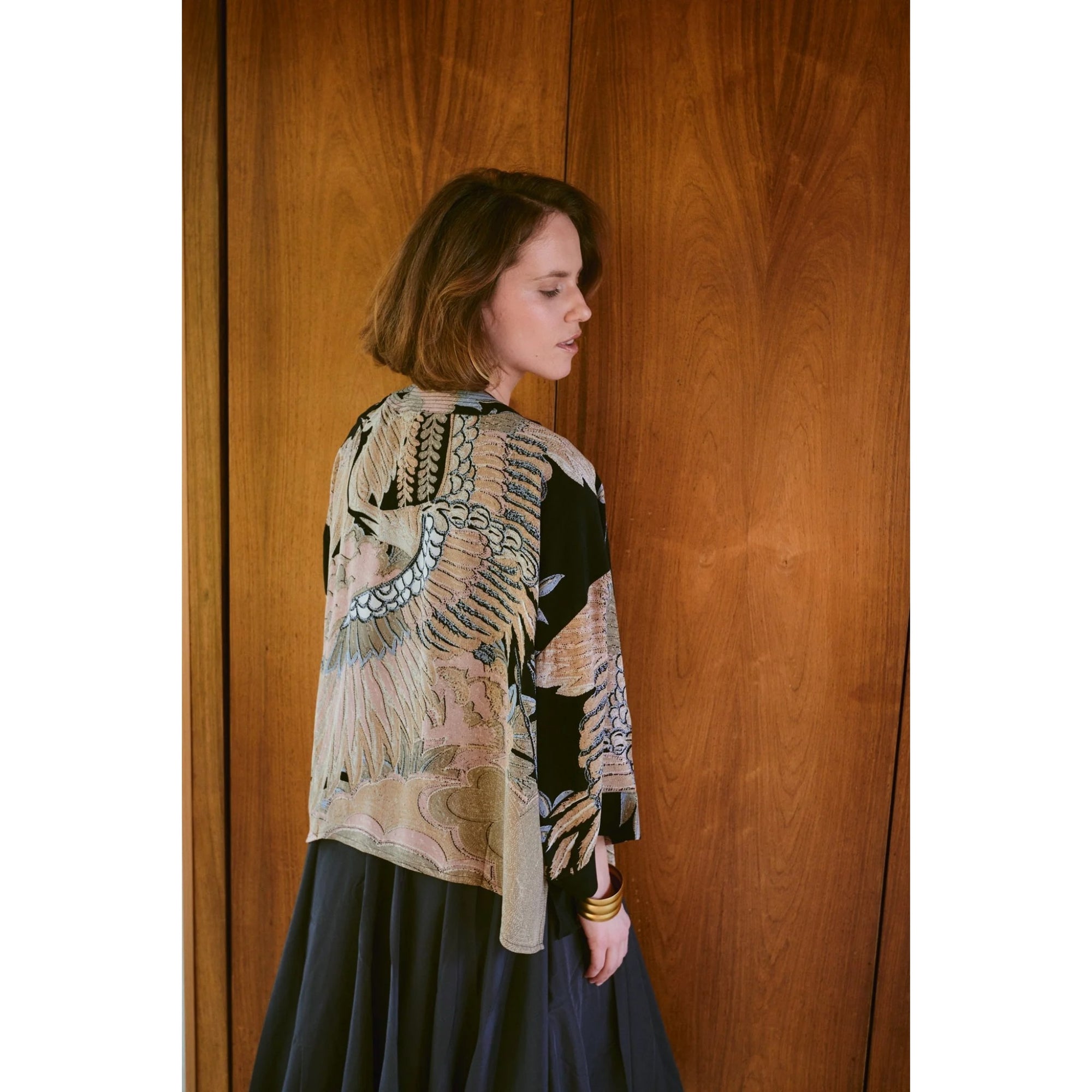 Woman wearing a patterned top and dark skirt against a wooden wall