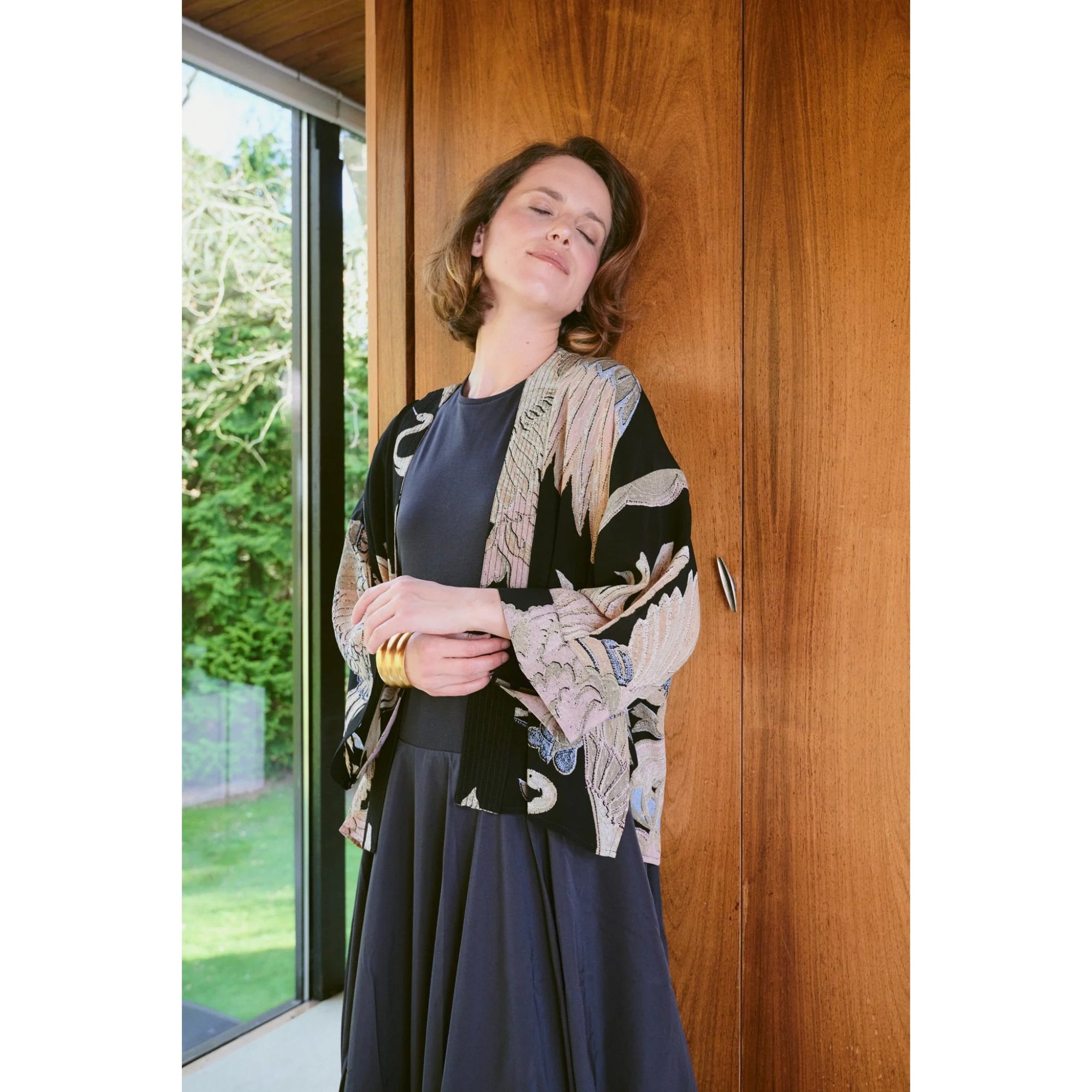 Woman wearing a patterned cardigan and dark dress standing indoors with a wooden wall and glass door in the background.