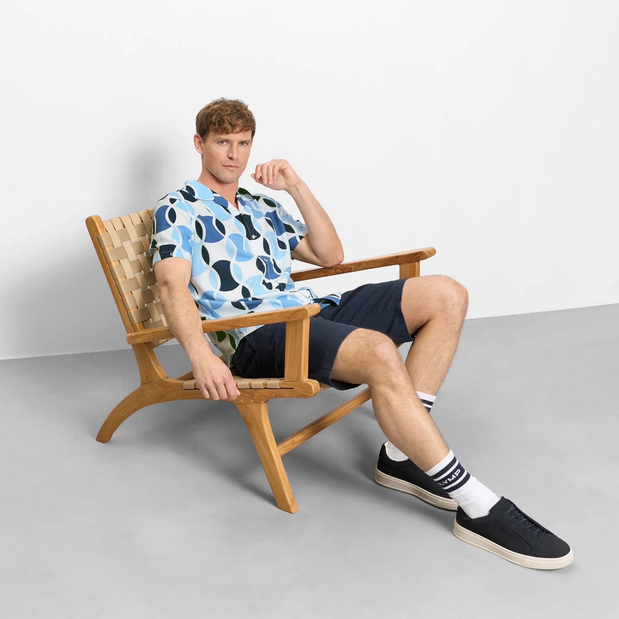 Man sitting on a wooden chair wearing a patterned shirt and shorts, with a plain background