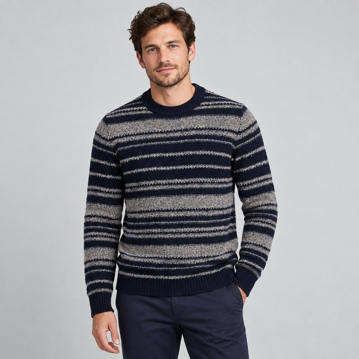 A man wearing a navy and gray striped knit sweater with dark navy trousers, standing against a light gray studio background.