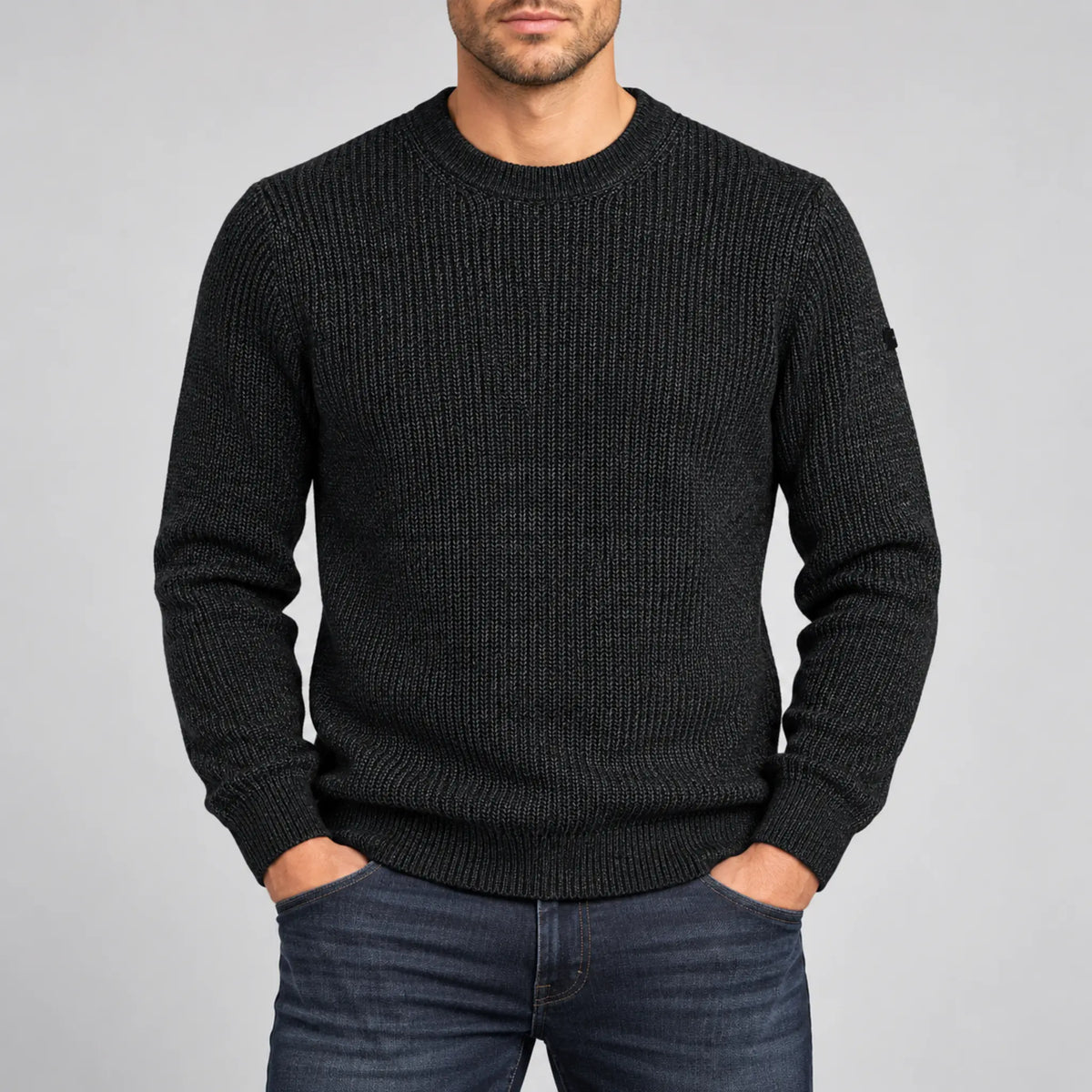 A man wearing a dark green ribbed knit crewneck sweater and dark blue jeans, standing against a light gray studio background.