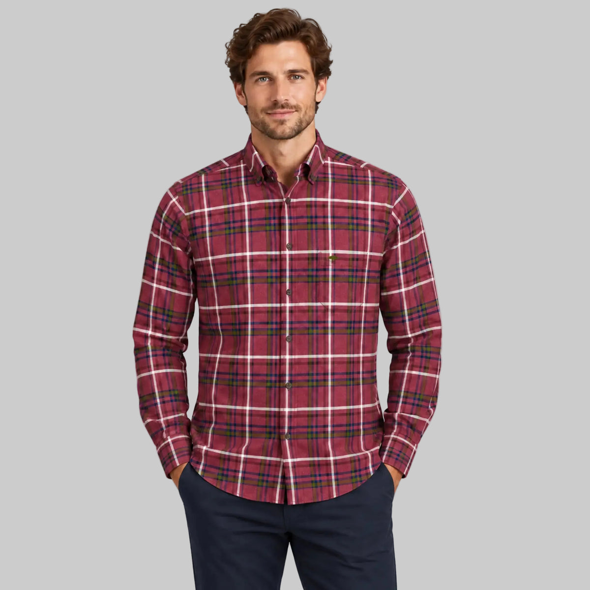 A man wearing a red and green plaid long-sleeve button-down shirt with white stripes, paired with navy pants, standing against a light gray studio background.