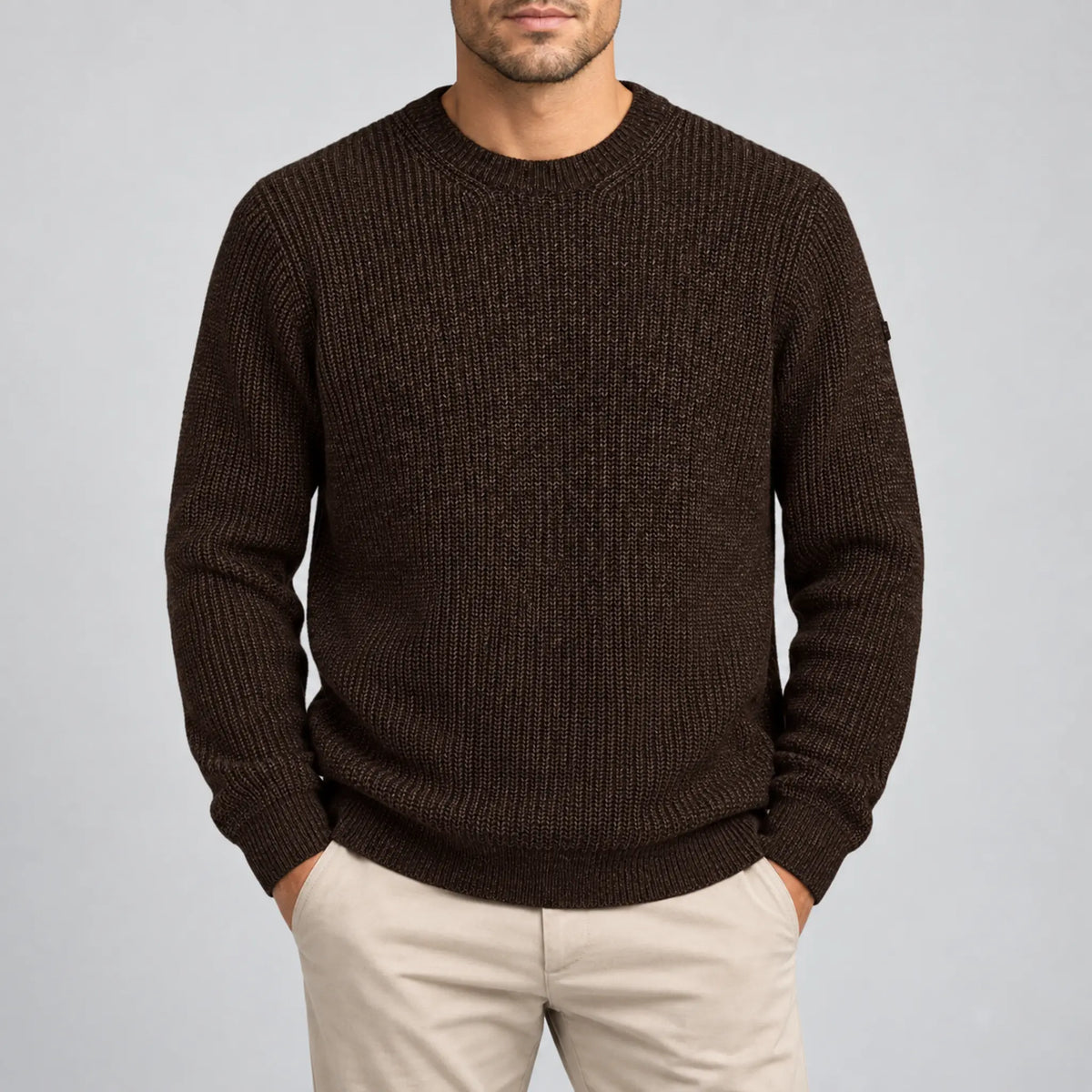 A man wearing a dark brown ribbed knit crewneck sweater and beige trousers, standing against a light gray studio background.