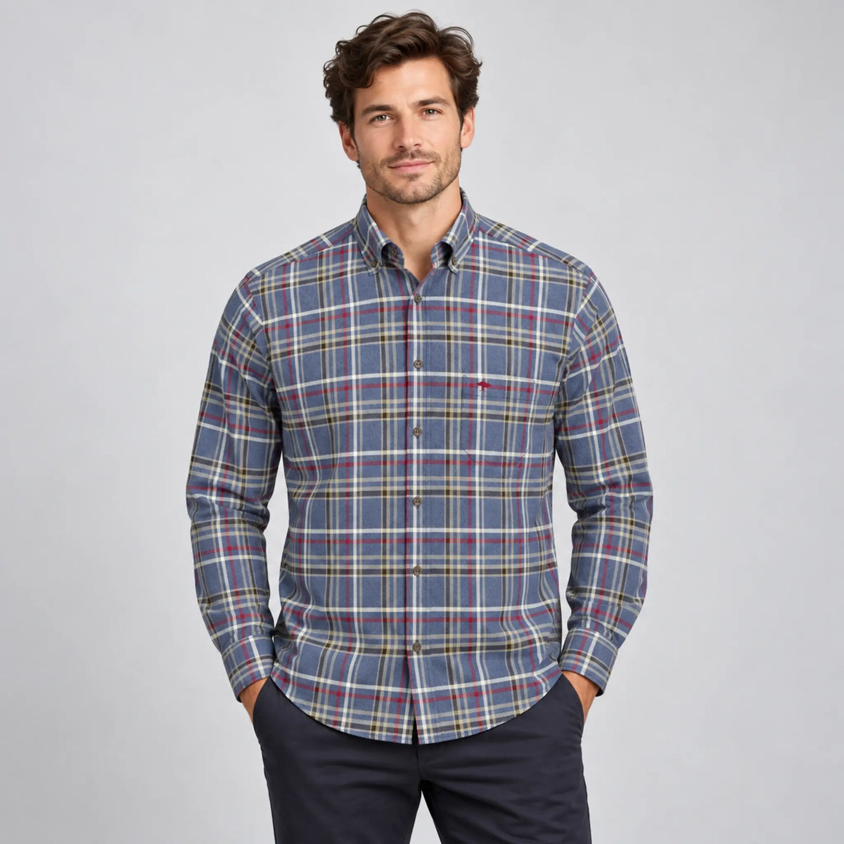 A man wearing a long-sleeve button-down plaid shirt in blue, red, and white, paired with dark trousers, standing against a light gray studio background.