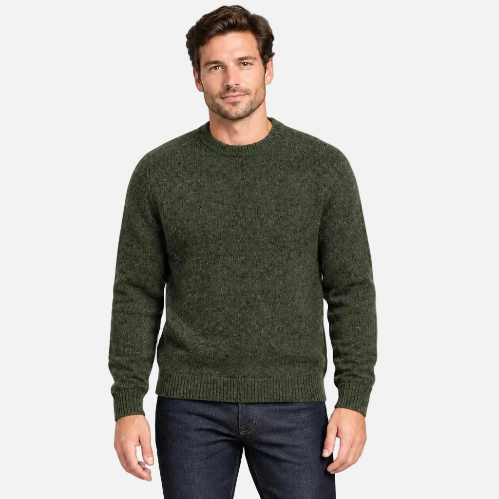 A man wearing a dark olive green knit crewneck sweater and dark blue jeans, standing against a solid light gray background.