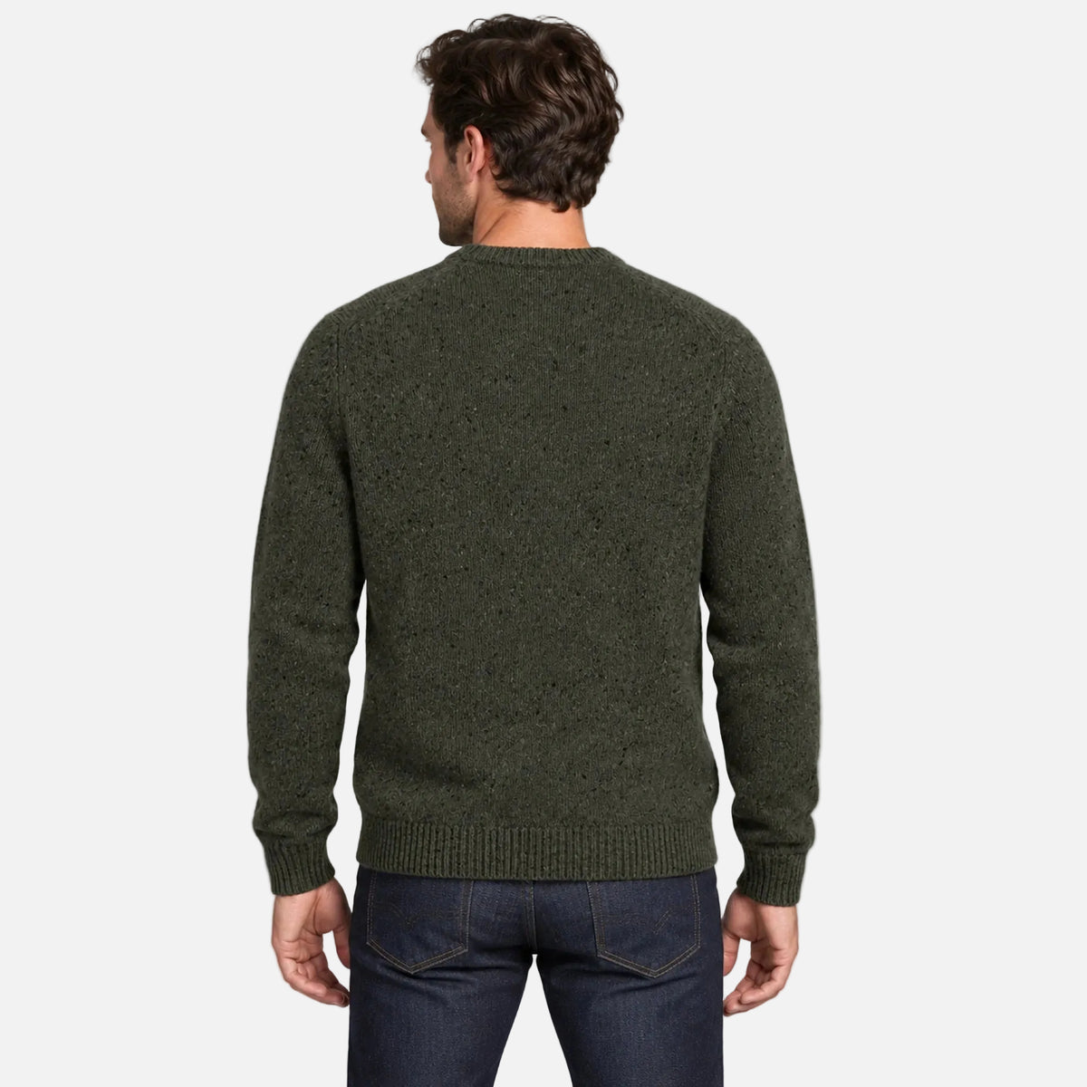 A man viewed from the back wearing an olive green knit sweater and dark blue jeans against a light gray studio background.