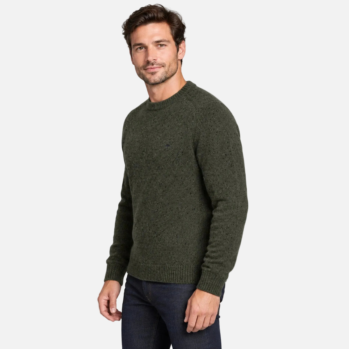 A man wearing a dark olive green knit crewneck sweater and dark blue jeans, standing against a solid light gray studio background.