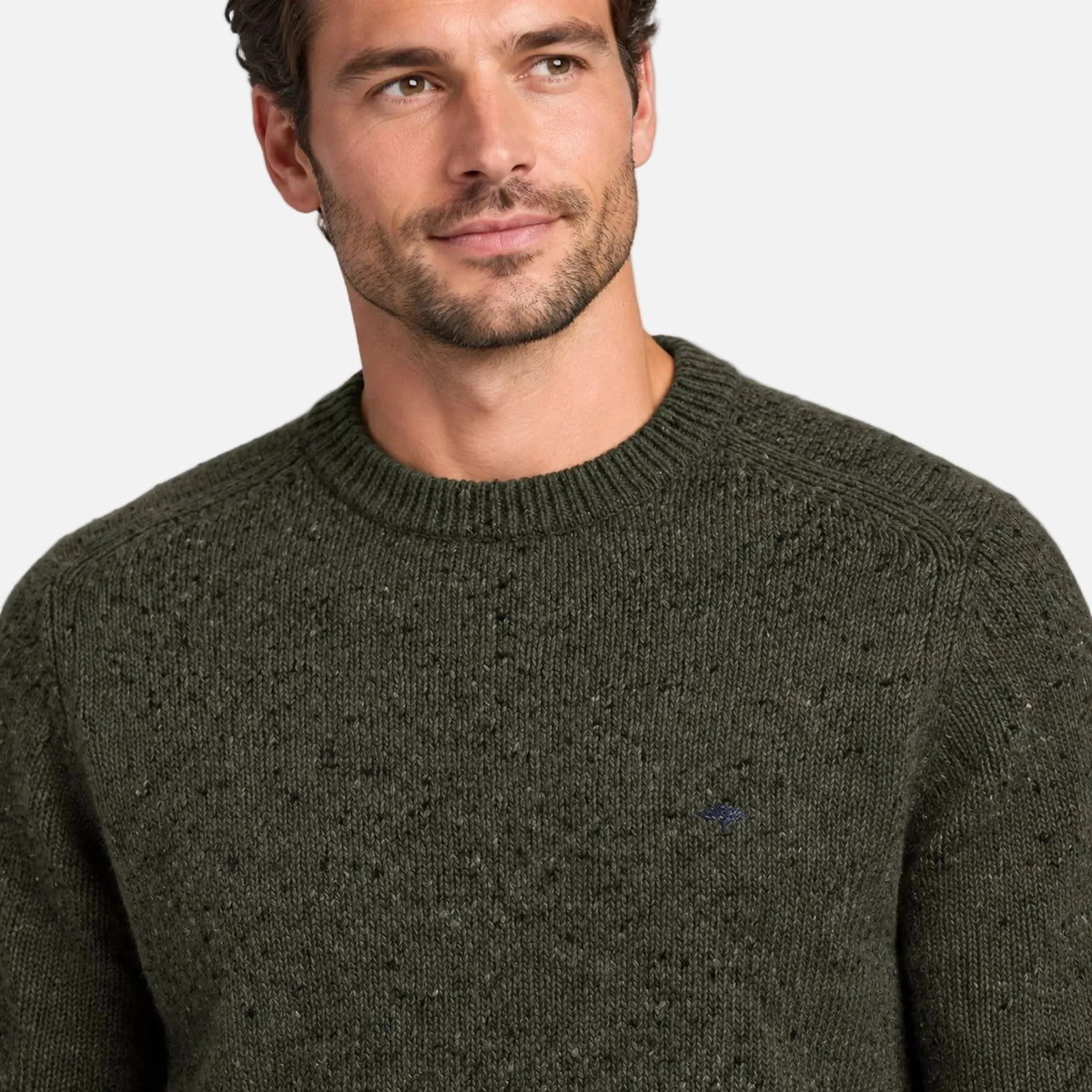 A close-up of a man wearing a dark olive green knit crewneck sweater against a light gray studio background.