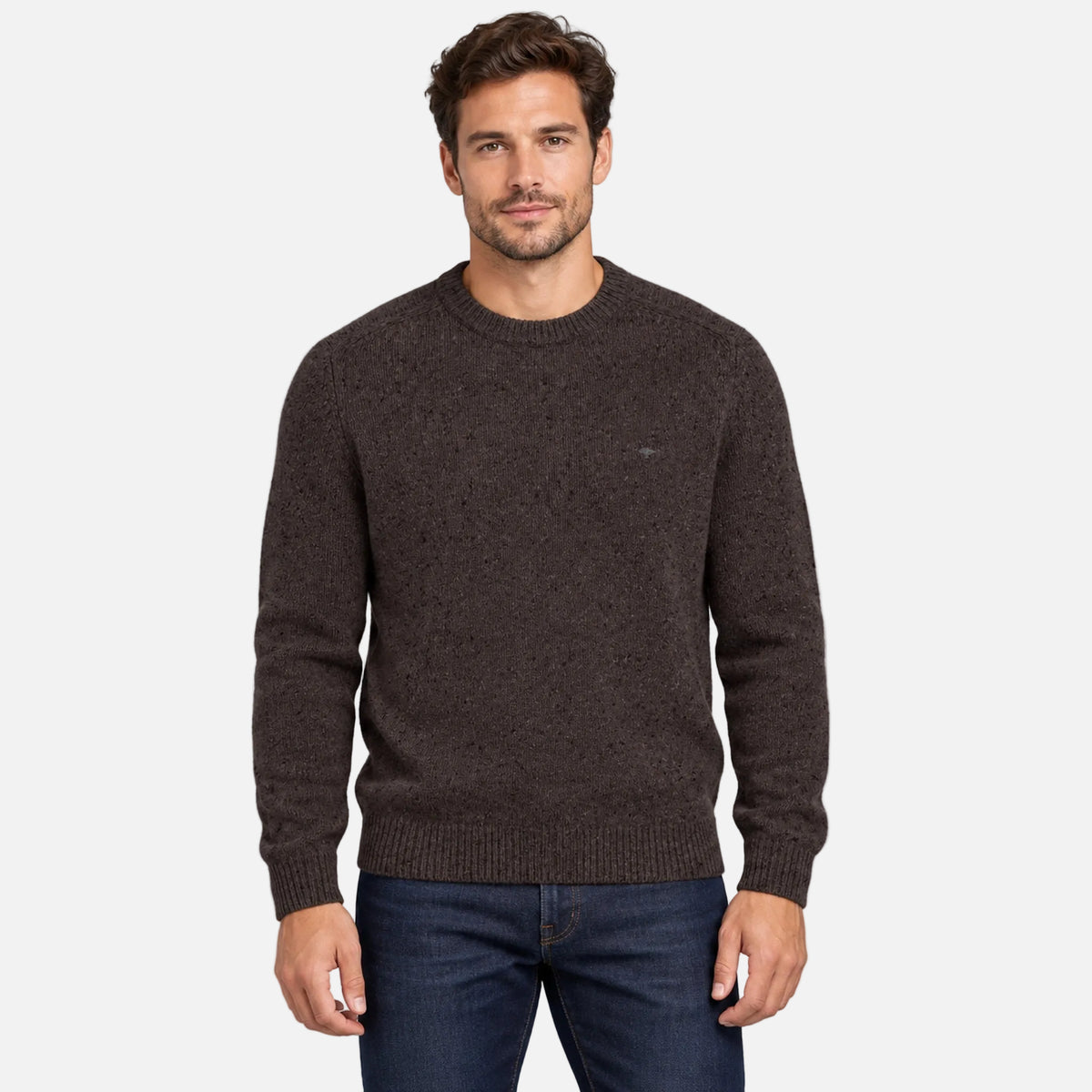 A man wearing a dark brown knit crewneck sweater and blue jeans, standing against a solid light gray background.