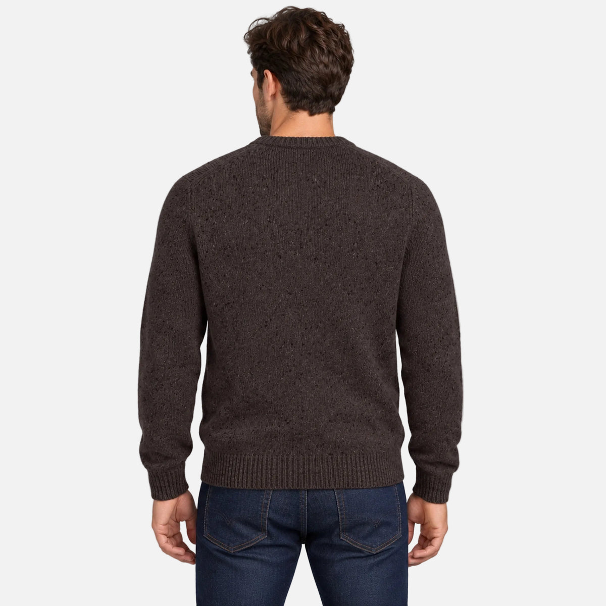 A man viewed from the back wearing a dark brown textured knit sweater and blue jeans, standing against a light gray studio background.