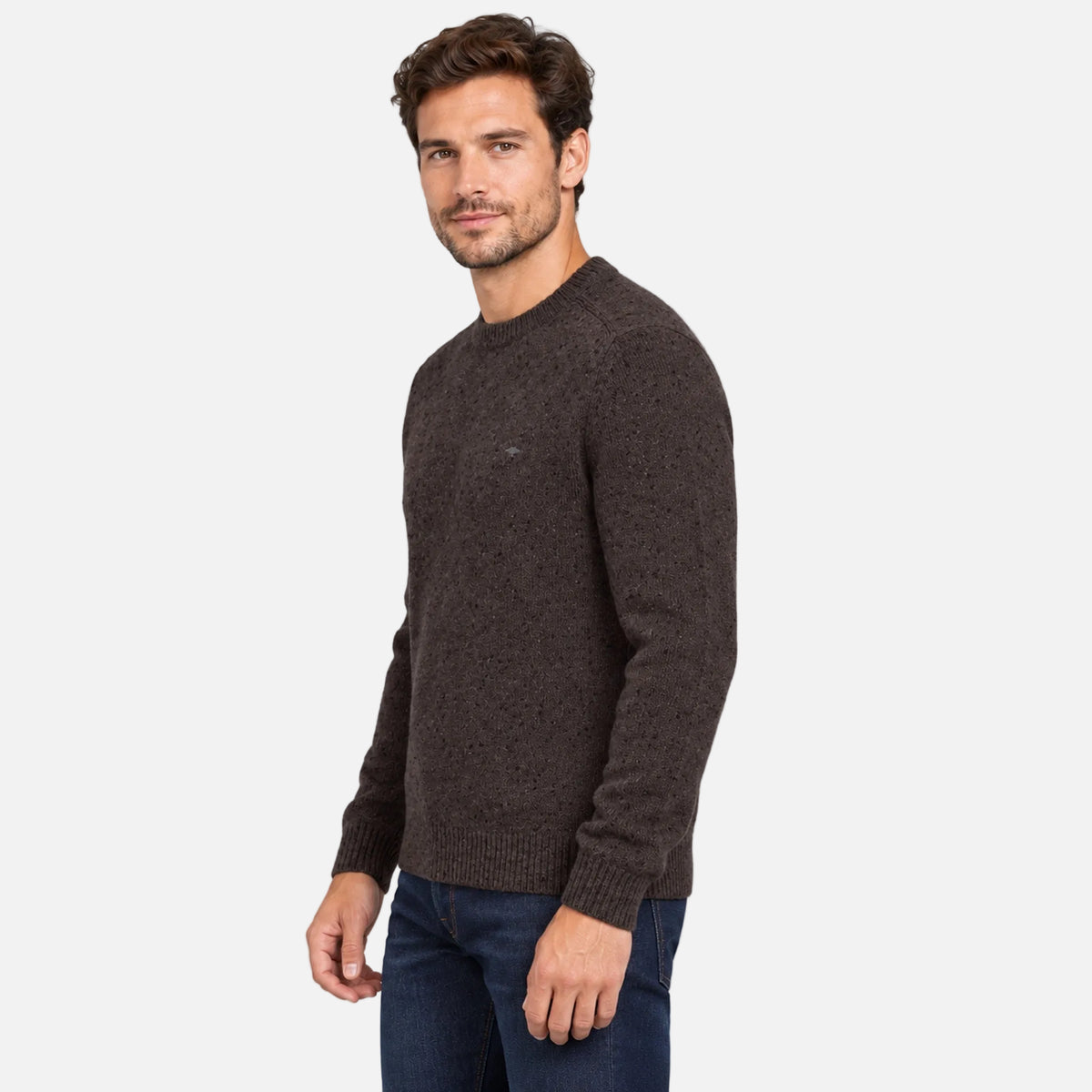 A man wearing a dark brown textured knit crewneck sweater and dark blue jeans, standing against a solid light gray background.