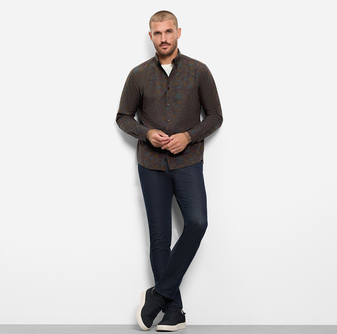 A man modeling a dark brown long-sleeve button-up shirt with a subtle geometric pattern, paired with dark blue jeans and black sneakers, standing against a light gray studio background.