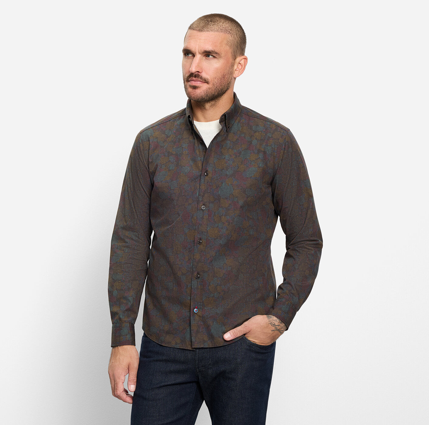 A man wearing a dark brown long-sleeve button-up shirt with a subtle blue and green paisley pattern, paired with dark blue jeans, standing against a light gray background.