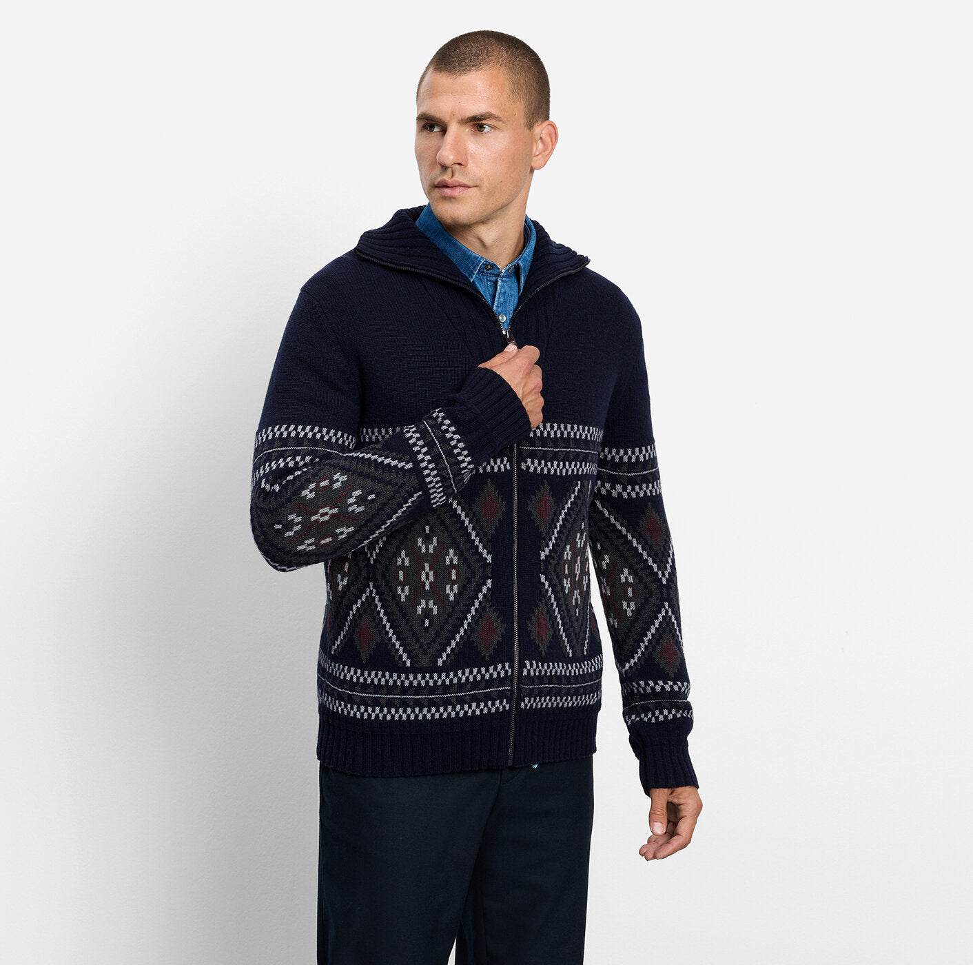 A man wearing a navy blue zip-up hoodie sweater with a white and burgundy geometric Fair Isle pattern, styled over a blue collared shirt and dark trousers, against a light gray studio background.