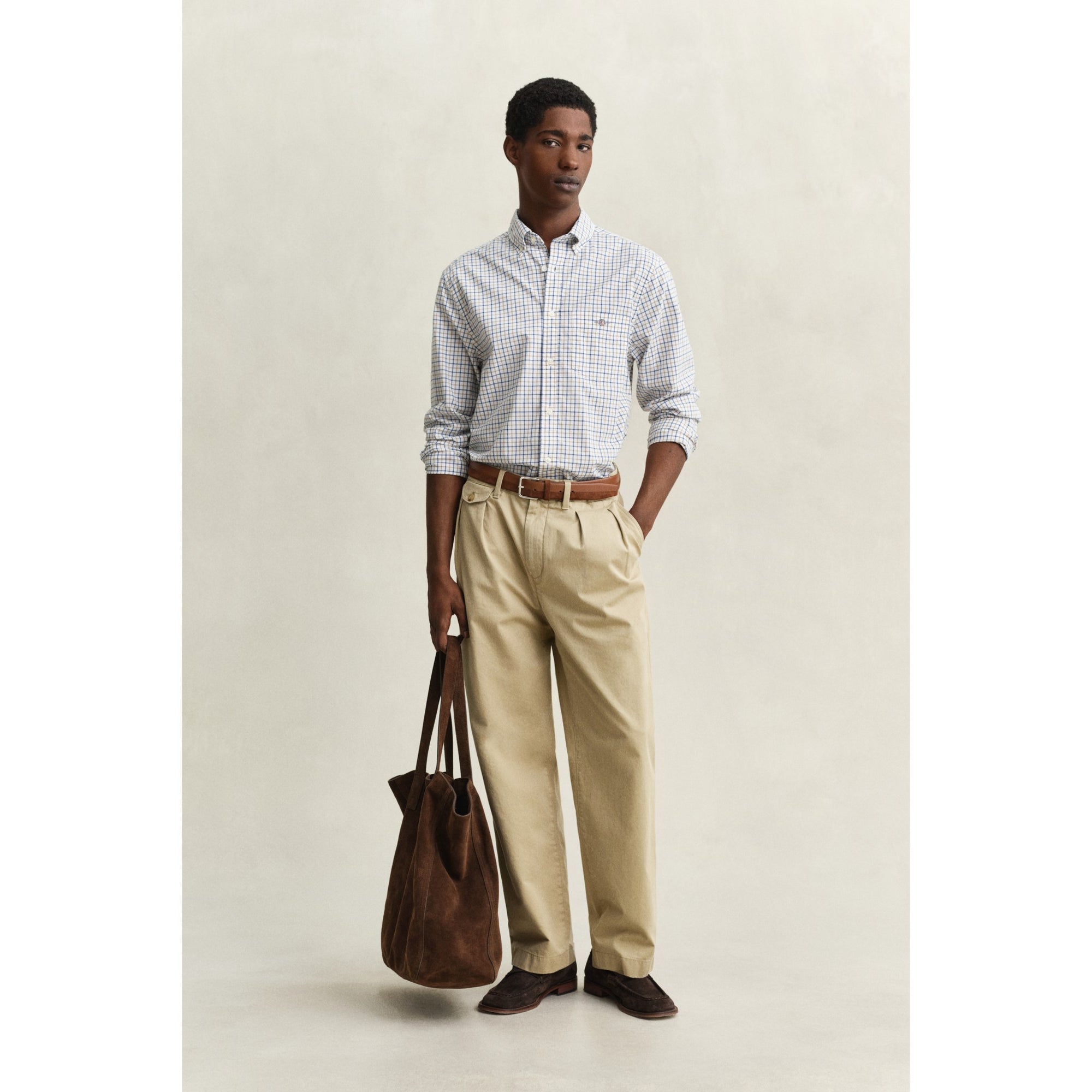 Man wearing a checkered shirt, beige pants, and brown shoes holding a leather bag against a plain background