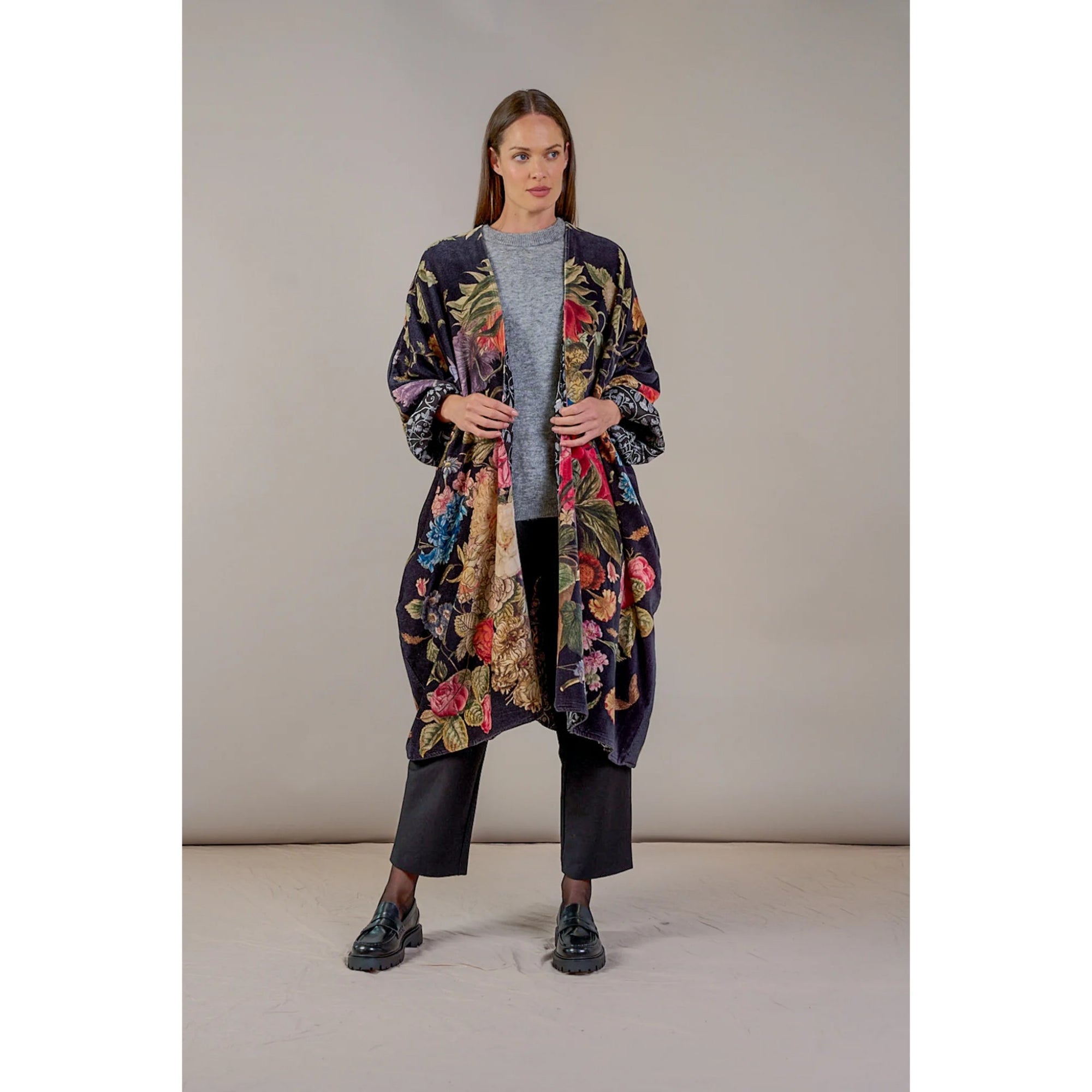 Woman wearing a long floral patterned coat over a gray sweater and black pants on a plain background