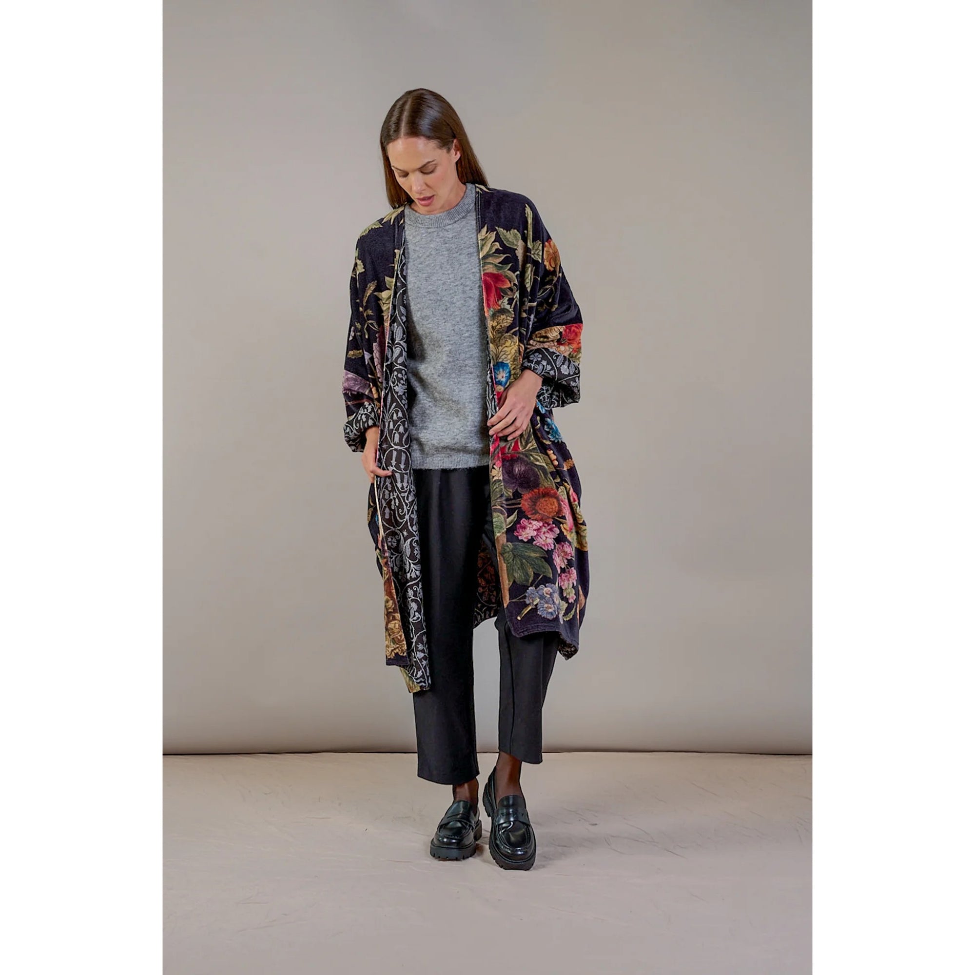 Woman wearing a long, floral-patterned coat over a gray sweater and black pants on a plain background.