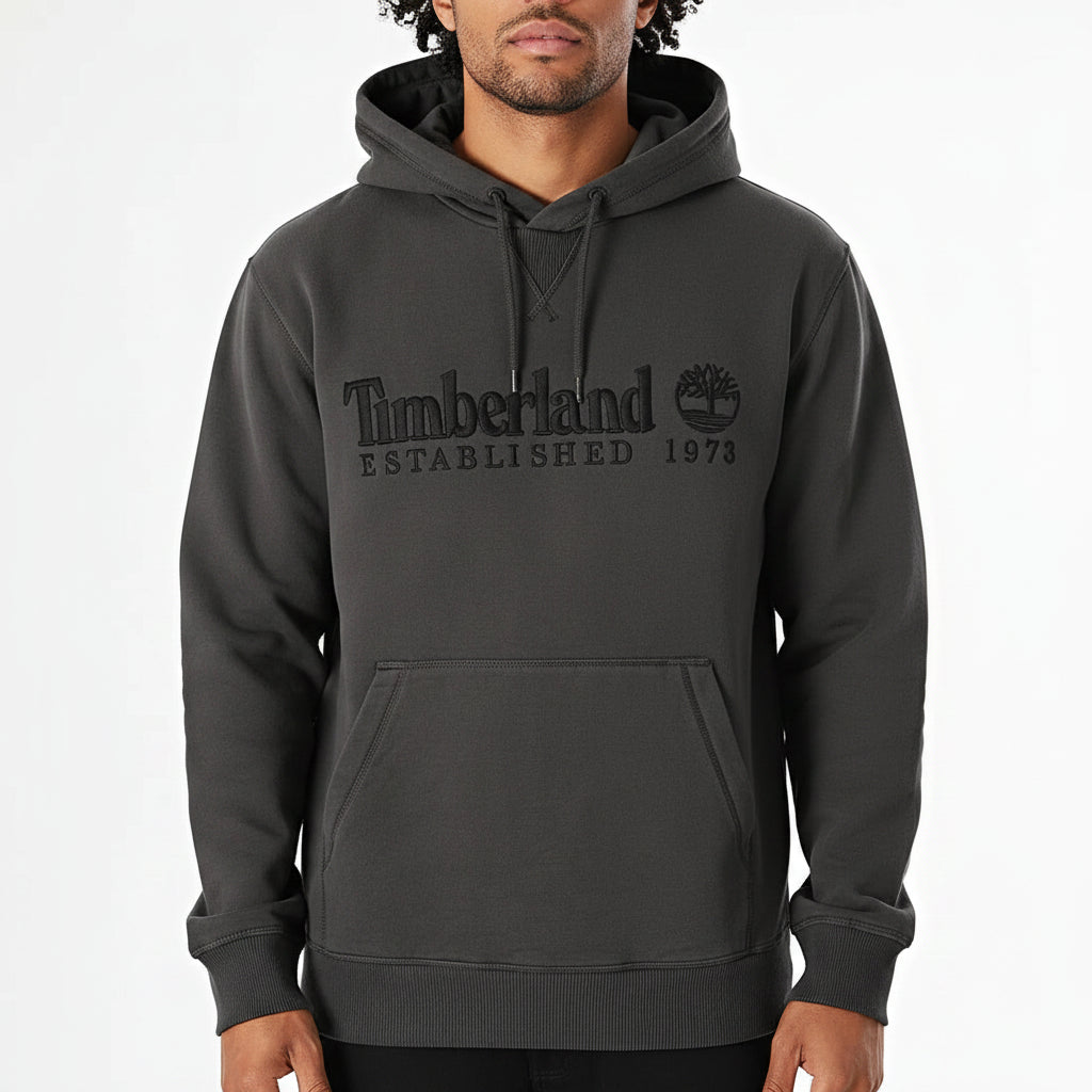 A man wearing a dark charcoal gray Timberland pullover hoodie with a tonal black embroidered logo on the chest, shown against a plain white background.