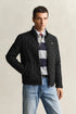 Man wearing a black quilted jacket over a patterned shirt with blue jeans on a plain background